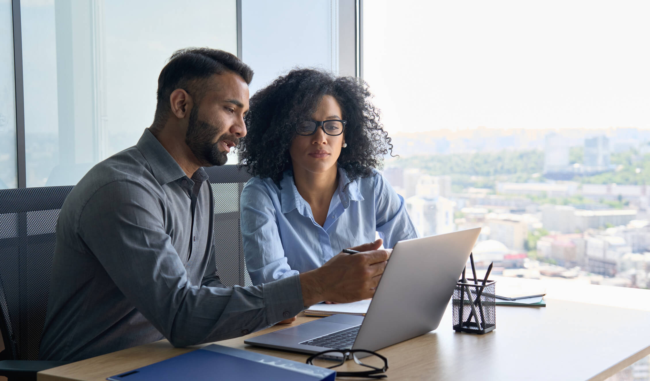 A man and woman sitting at a desk discussing what is on a laptop screen.